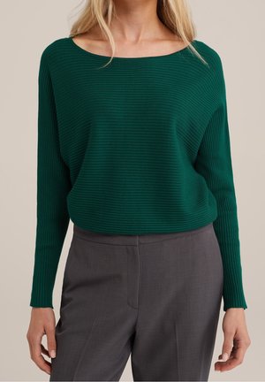 Femme portant un pull côtelé vert foncé à col bateau et un pantalon gris ajusté, debout les bras détendus le long du corps.