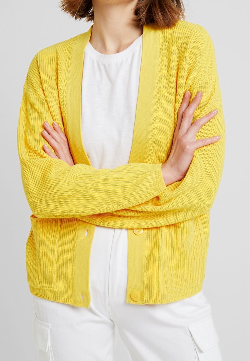 Personne portant un cardigan en maille jaune vif par-dessus une chemise blanche et un pantalon blanc, debout les bras croisés devant un fond uni.