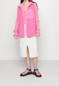 Gilet en polaire rose avec un corps texturé, une fermeture à boutons et des poches zippées, associé à une jupe midi à rayures blanches et des sandales colorées.