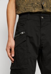 Pantaloni cargo negri, cu multiple buzunare, un buzunar lateral cu fermoar și o textură netedă. Se asortează cu o cămașă cu model.