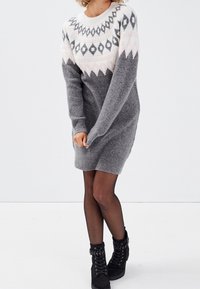 Robe-pull grise en tricot avec un col rond, décorée de motifs géométriques noirs et roses, dotée de manches longues et de poignets côtelés.