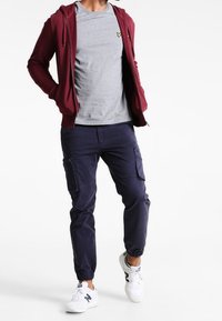 Man som bär en vinröd zip-up hoodie, grå t-shirt, marinblå cargo-joggers och vita sneakers, går med händerna i fickorna.