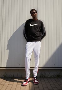 Man wearing black Nike sweatshirt, white Nike track pants, red and white Nike sneakers, and sunglasses standing against a light gray wall.