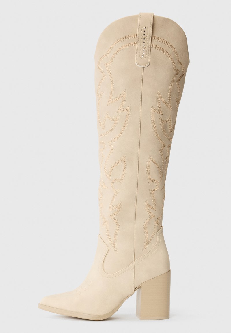 Botte de cowboy à talon haut beige en microsuède avec des détails brodés sur la tige et un nez carré. Présente un talon large.