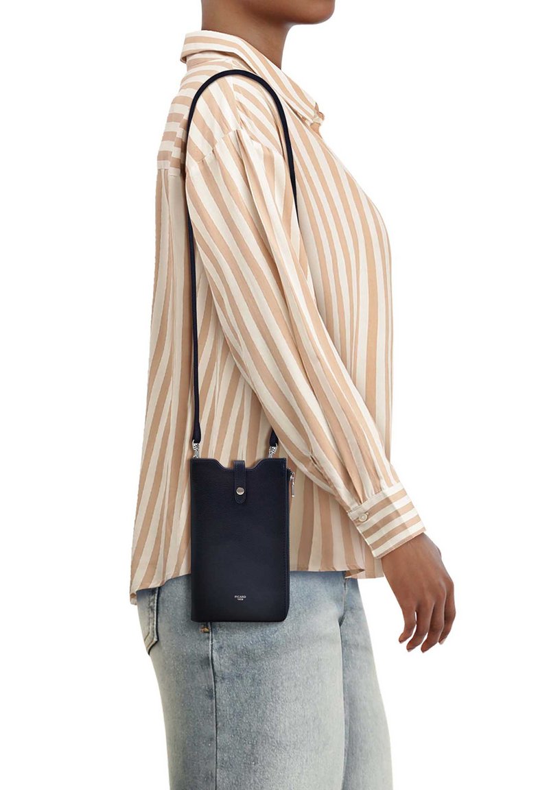 Personne portant une chemise à rayures beige et blanc et un jean clair, portant un petit sac bandoulière noir avec une sangle sur l'épaule.