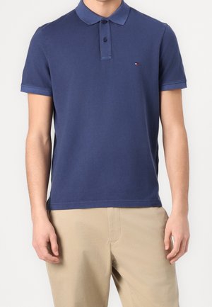 Mężczyzna w granatowej koszulce polo z krótkim rękawem i małym logo na piersi oraz w beżowych spodniach, stojący na białym tle.