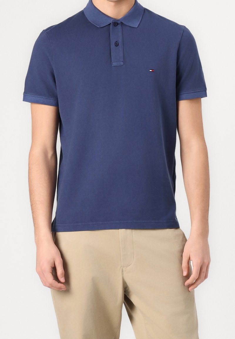 Homme portant un polo bleu marine à manches courtes avec un petit logo sur la poitrine et un pantalon beige, debout devant un fond blanc.