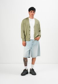 Homem de camisola canelada verde-clara, t-shirt branca, calções de ganga claros até ao joelho, meias brancas e ténis pretos, de pé contra um fundo branco.