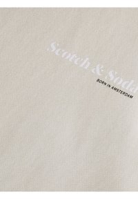 Tejido de algodón en beige con un tejido texturizado. Presenta texto impreso en blanco "Scotch & Soda" y "BORN IN AMSTERDAM" en negro.