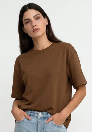 Kobieta o długich ciemnych włosach, ubrana w luźną brązową koszulę z krótkim rękawem wpuszczoną w jasnobrązowe dżinsy, z rękami w kieszeniach.