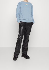 Persona che indossa una felpa azzurra chiara con il logo "Hugo", pantaloni di pelle nera e scarpe nere chunky, in piedi con le mani in tasca.