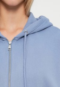 Svijetloplava zip-up dukserica s teksturom tkanine, vezicama i srebrnim dijelovima na patent-zatvaraču. Približeni pogled na izrez.