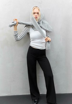 Giovane donna con pantaloni neri e maglietta a righe a maniche lunghe che avvolge un foulard grigio attorno alla testa, in piedi di fronte a un muro grigio uniforme.