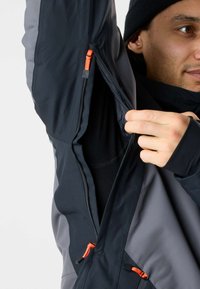 Homme portant une veste noire et grise dézippant une fermeture éclair de ventilation sous les bras avec des curseurs orange.