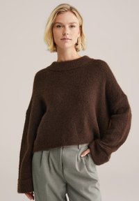 Pulover scurt de culoare maro, cu o textură tricotată lejeră și mâneci largi, asortat cu pantaloni gri deschis. Aspect moale și confortabil.