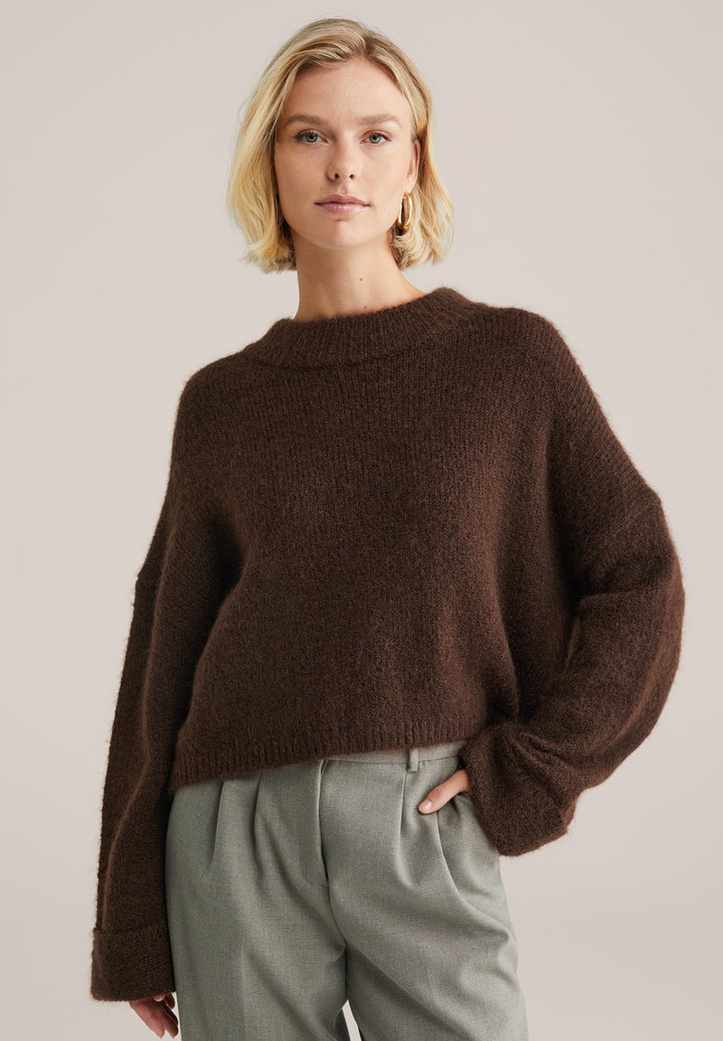 Hnědý zkrácený svetr s volnou pletenou strukturou a širokými rukávy, spárovaný se světle šedými kalhotami. Jemný, útulný vzhled.