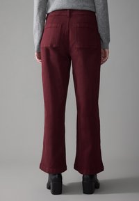 Persona vistiendo pantalones burdeos de tiro alto y pierna recta con bolsillos traseros, una camiseta de manga larga gris y botas negras de tacón en el tobillo, de pie sobre un fondo gris.