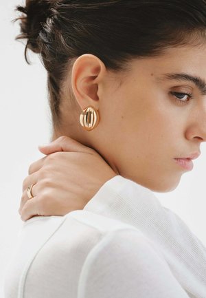 Pendientes en tono dorado con un diseño escultórico que presenta líneas curvas y un acabado brillante, llevados por una persona con una blusa blanca y un peinado elegante.