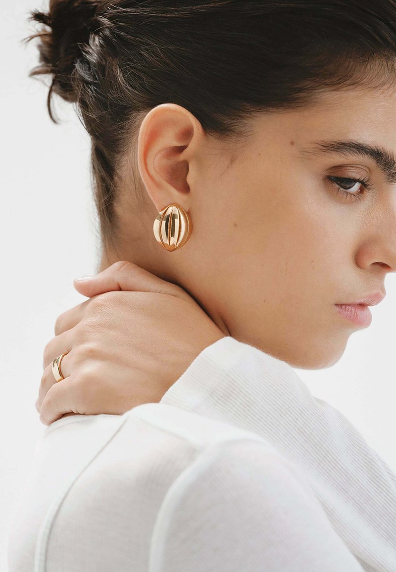 Pendientes en tono dorado con un diseño escultórico que presenta líneas curvas y un acabado brillante, llevados por una persona con una blusa blanca y un peinado elegante.