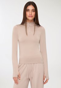 Beżowa, długo rękawowa bluzka z wysokim dekoltem, wykonana z gładkiego materiału. Posiada zamek z przodu, dopasowany krój oraz otwory na kciuki w mankietach.