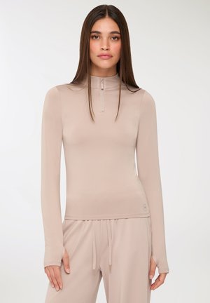 Top a maniche lunghe e collo alto color beige, realizzato in tessuto morbido. Presenta una zip frontale, una vestibilità slim e aperture per il pollice nei polsini delle maniche.