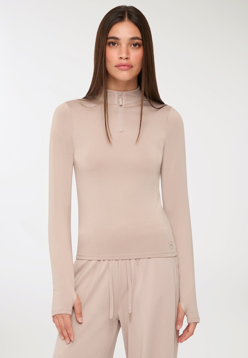 Beżowa, długo rękawowa bluzka z wysokim dekoltem, wykonana z gładkiego materiału. Posiada zamek z przodu, dopasowany krój oraz otwory na kciuki w mankietach.
