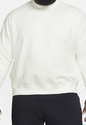 Mężczyzna w jednolitej białej bluzie Nike z długim rękawem i małym białym logo na piersi, w czarnych spodniach, stojący z rękami przy bokach.