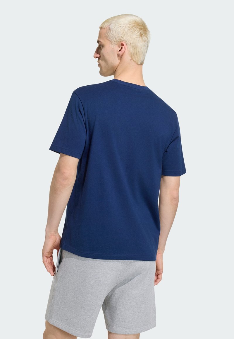 Homme blond portant un t-shirt bleu marine uni à manches courtes et un short gris clair, debout avec le dos partiellement tourné.