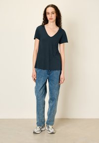 Temnozeleno majico s V-izrezom in kratkimi rokavi, v kombinaciji s svetloplavimi ohlapnimi kavbojkami in sivo obutvijo z modrimi detajli.