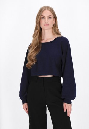 Marineblaue, verkürzte Sweater mit langen, puffigen Ärmeln und einem lässigen Ausschnitt, kombiniert mit hoch taillierten schwarzen Hosen. Glatte Strickstruktur.