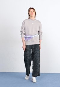 Lys grå hettegenser med en liten logo, svarte bukser med blomsterprint, og en lilla rompetaske. Hvite sneakers fullfører antrekket.