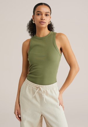 Noor neiu lokkis juustega, kandes kuldseid rõngakõrvarõngaid, oliivrohelist varrukateta topi ja kreemja nööriga pükse ühtlasel taustal.