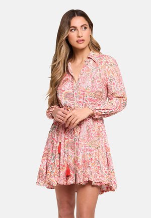 Mujer con el pelo largo que lleva un vestido de manga larga con estampado de paisley en rosa y naranja y un dobladillo con volantes, mirando hacia un lado.