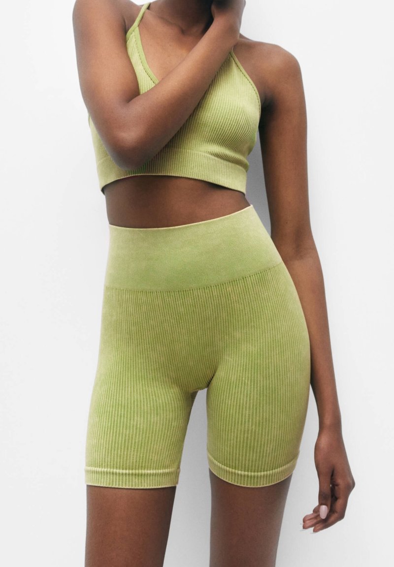 Femme portant un crop top côtelé vert clair et un short de cycliste taille haute, debout devant un fond blanc.