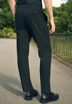 Person in einer schwarzen Hose und schwarzen Lederloafers steht auf einem gepflasterten Weg in der Nähe eines Metallzauns und von Grünpflanzen.