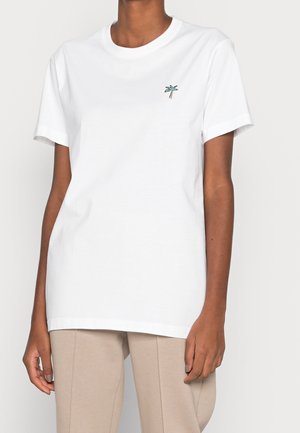 Person wearing a plain white short-sleeve t-shirt with a small green palm tree embroidered on the left chest, paired with beige pants.