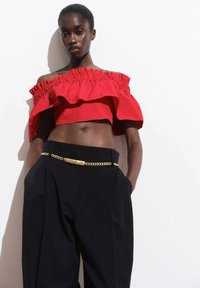 Top corto rosso off-the-shoulder con dettagli a ruches, abbinato a pantaloni neri a vita alta e una cintura in catena dorata. Tessuto dalla consistenza morbida.