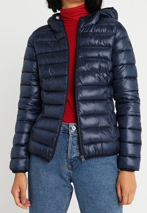 Femme portant une veste matelassée bleu marine avec capuche, un col roulé rouge et un jean bleu, debout les mains détendues le long du corps.