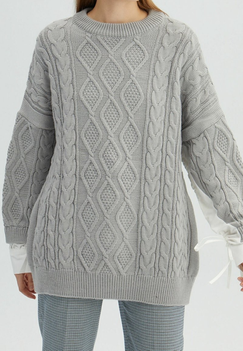 Grijze kabelgebreide trui met textuur van diamant- en vlechtpatronen, oversized pasvorm en geribbelde manchetten. Gelaagd met een wit overhemd eronder.