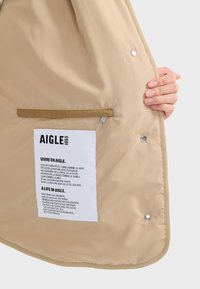 Beige Oberbekleidung mit glatter Textur, ausgestattet mit einem weißen Etikett mit schwarzem Text und einer Reißverschlusstasche, die mit Druckknöpfen gesichert ist.
