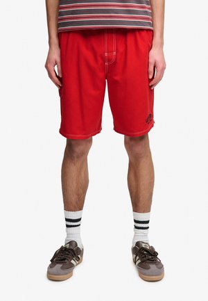 Mężczyzna w czerwonych szortach z białymi szwami, białych skarpetkach z dwoma czarnymi paskami oraz brązowych sportowych butach, stojący na jednolitym białym tle.