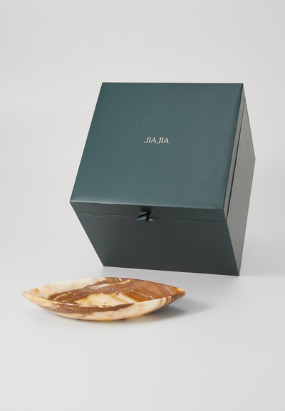 Dunkelgrünes quadratisches Kästchen mit goldener "JIA JIA"-Aufschrift neben einem ovalen, marmorierten braunen und cremefarbenen dekorativen Teller auf einer weißen Oberfläche.