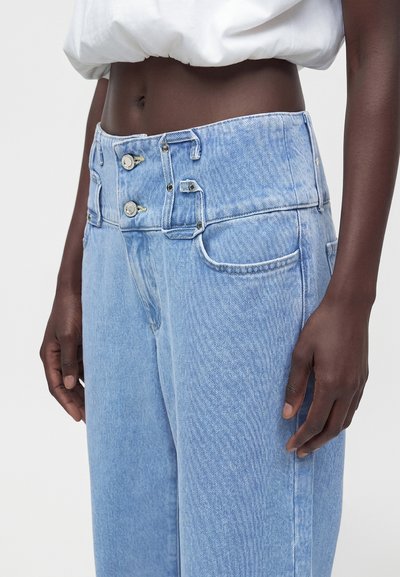 Taille et hanches d’une personne portant un jean bleu clair taille haute à double boutonnière et un top court blanc.