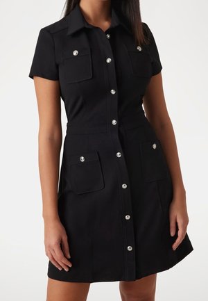 Woman wearing a black short-sleeve button-down dress with four flap pockets, silver buttons, and a collar against a white background.