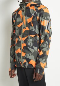 Giacca con cappuccio caratterizzata da un motivo di camouflage geometrico nei colori verde, nero e arancione. Include zip frontale, tasche laterali e dettagli riflettenti.