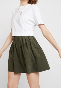 T-shirt blanc court à manches courtes associé à une jupe plissée vert olive foncé. La jupe a une texture lisse et des détails de couture subtils.