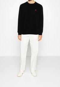 Sudadera negra con cuello redondo y pequeño logo rojo en el pecho, combinada con pantalones blancos y zapatos de color claro. Textura suave.