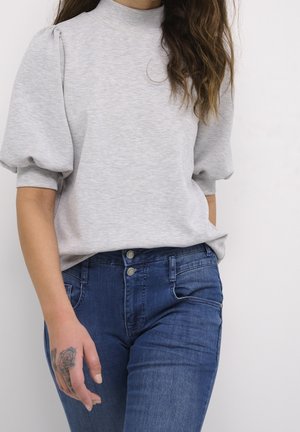 Femme portant un haut gris clair à manches bouffantes et un jean bleu taille haute avec un tatouage de rose sur le dos de sa main gauche.