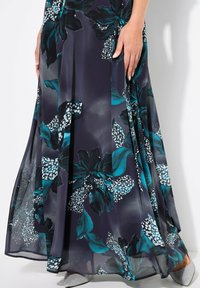 Maxikleid aus transparenter Stoff mit navyfarbenem Grund, das große dunkelblau-grüne Blumenmuster und weiße Pünktchen aufweist. Weiche Falten und ein fließender Saum.