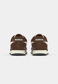 Par cipela Reebok smeđe i bijele boje prikazanih straga s vidljivim logotipom na peti i crnim gumenim potplatima na jednostavnoj bijeloj pozadini.